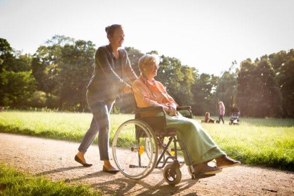 一个四肢瘫痪的人坐在轮椅上，在看护者的陪同下在家附近的公园散步