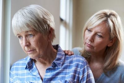 senior woman with blank facial expression looking out window while her daughter puts hand on her shoulder