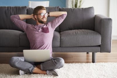 man sitting on living room floor with laptop, smiling and putting hands behind head