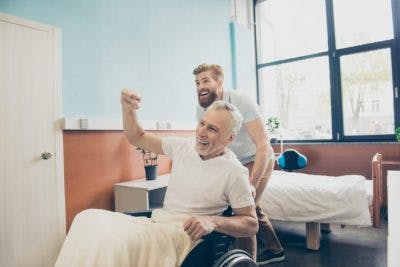 adult grandson wheeling grandfather out of hospital room while grandfather cheers