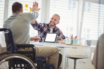 man sitting at work desk high-fiving coworker who is in a wheelchair