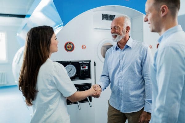 senior man shaking hands with nurse in front of hyperbaric oxygen therapy chamber that he will use to treat his brain injury