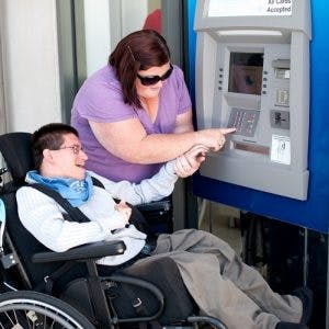 A young man with severe cerebral palsy in a wheelchair being assisted to use an ATM.
