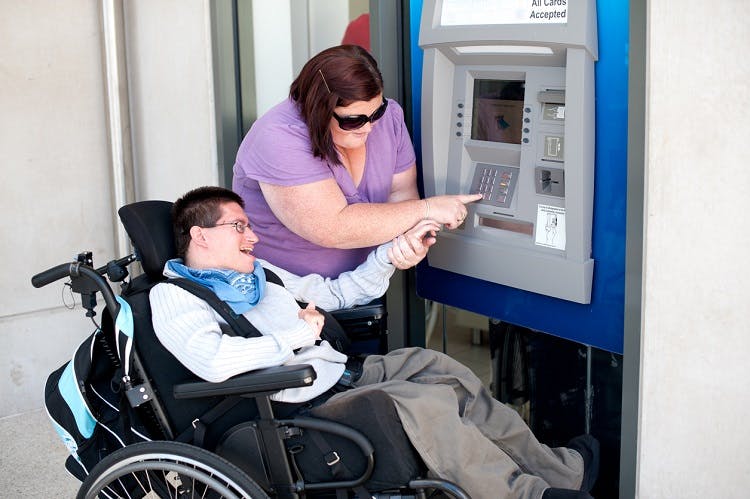 A young man with severe cerebral palsy in a wheelchair being assisted to use an ATM.