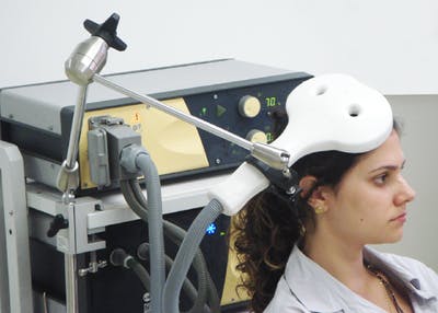 Woman seated in clinical chair with huge magnet resting above her head for rTMS stroke recovery treatment