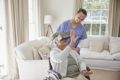 stroke survivor in wheelchair with virtual reality set over her eyes and her arms lifted as if she's seeing something, while her caregiver looks down at her smiling